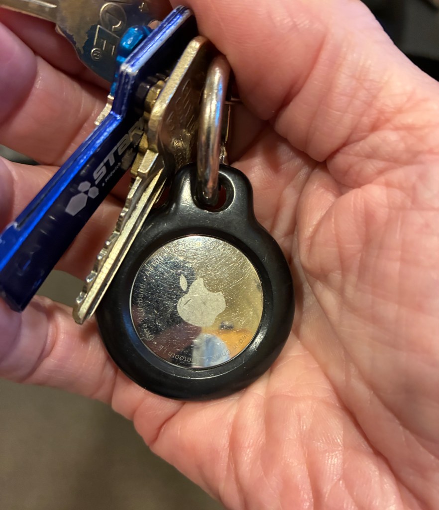 A person's hand holding a set of keys with a metallic Apple AirTag attached.