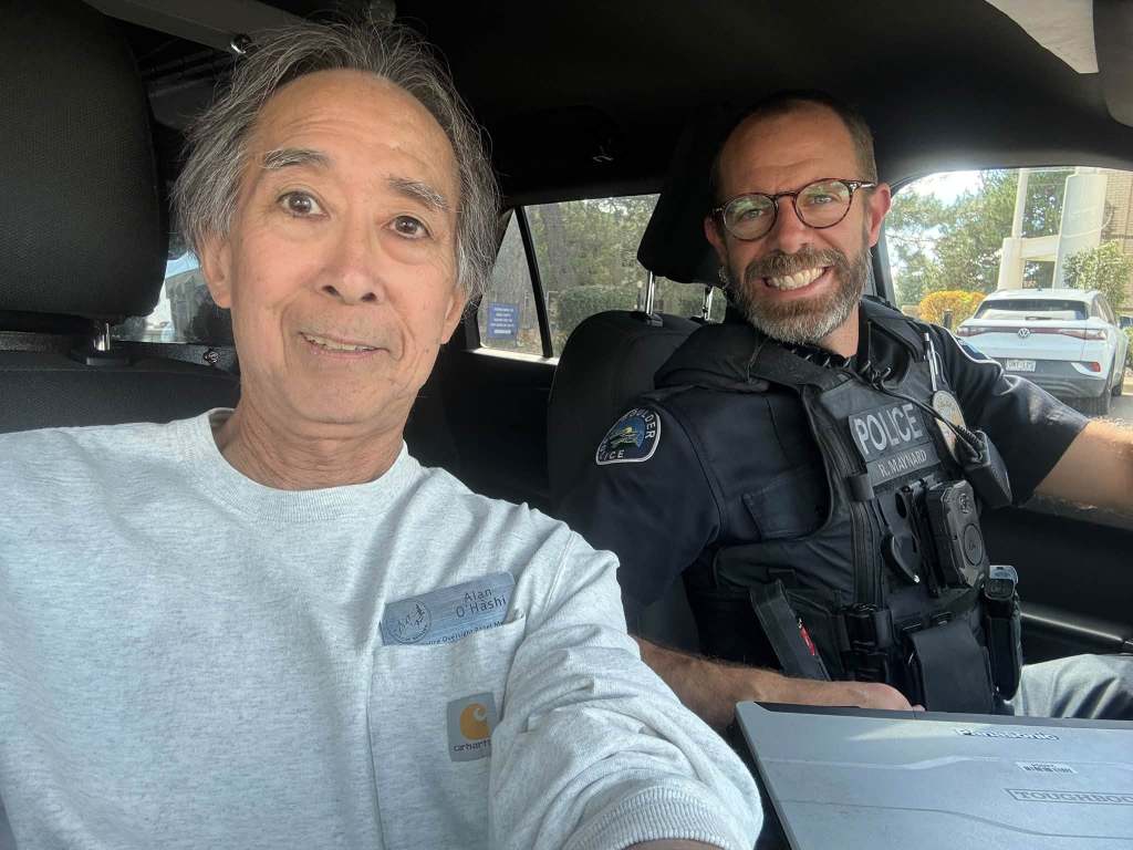 A selfie of two men inside a police vehicle; one man is wearing a gray sweatshirt and smiling, while the police officer, wearing a uniform and glasses, smiles next to him.
