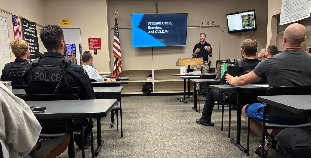 A police training session in a classroom setting, with officers seated at tables and a speaker presenting concepts related to probable cause and searches on a screen.