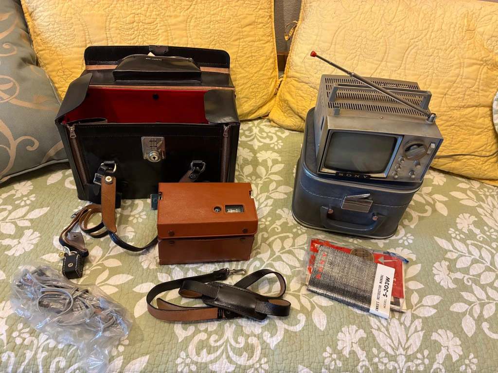 A collection of vintage electronics including a black leather camera bag, a brown camera, a small vintage Sony television with a retractable antenna, and additional accessories laid on a floral patterned fabric.