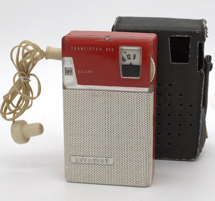 A vintage transistor radio, red and silver in color, with a volume control and an earphone connected, next to a black carrying case.