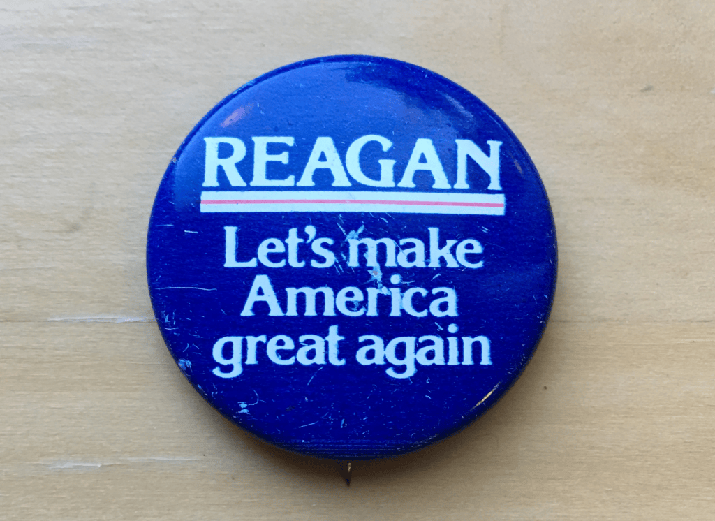 A blue campaign button featuring the word 'REAGAN' in bold white letters, with the slogan 'Let's make America great again' underneath.