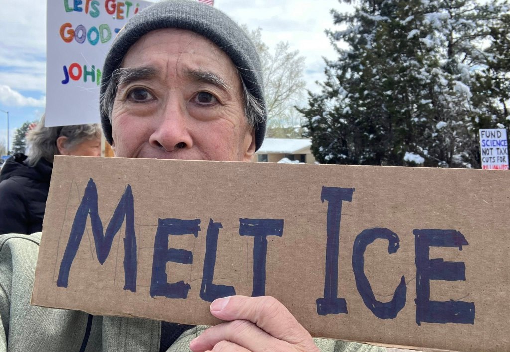 A person wearing a gray beanie holds a cardboard sign that reads 'Melt Ice' at a protest, with other protesters and signs visible in the background.