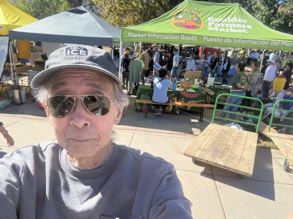 A person taking a selfie at the Boulder Farmers Market, with information booths and a crowd visible in the background. The person wears sunglasses and a cap, and there are colorful market tents set up.