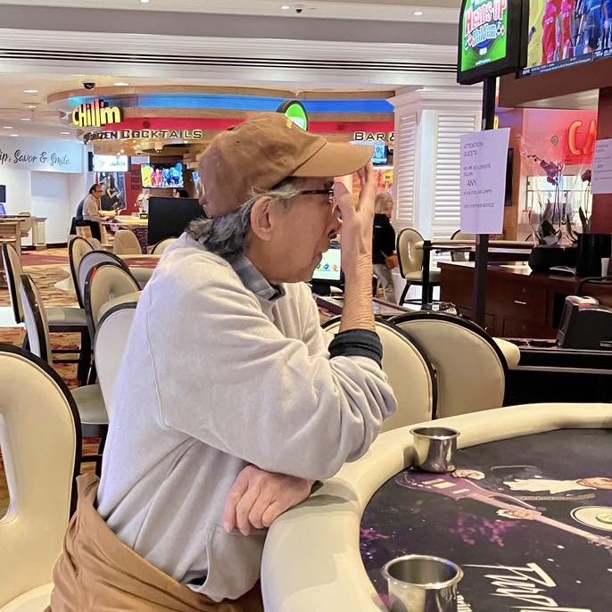 A man sitting at a casino table with a concerned expression, wearing a baseball cap and a light sweater, in a vibrant casino environment with digital screens in the background.