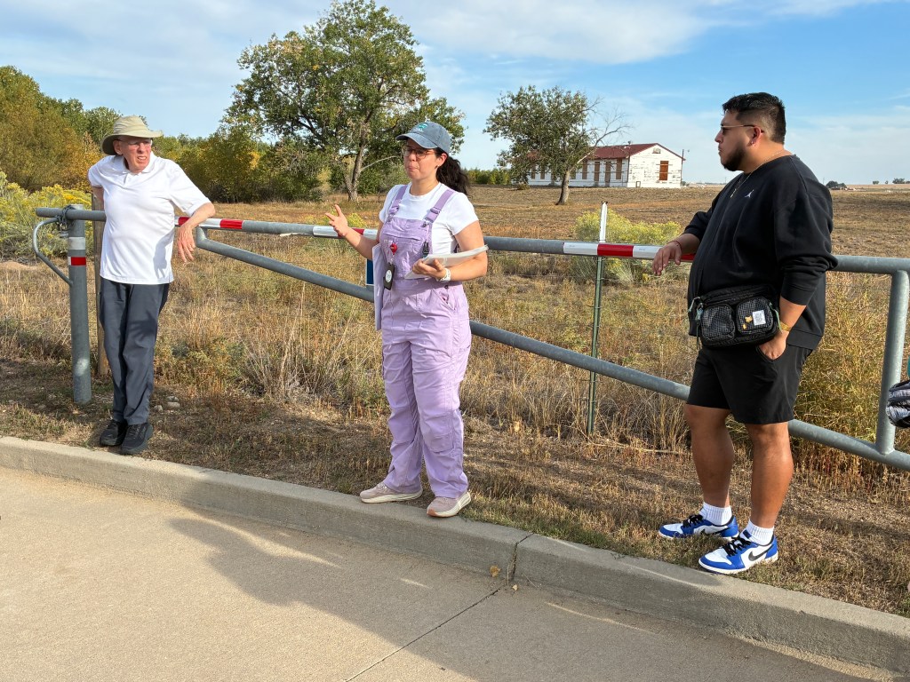 Three individuals engaged in conversation by a gate outdoors, with a grassy area and a building in the background.