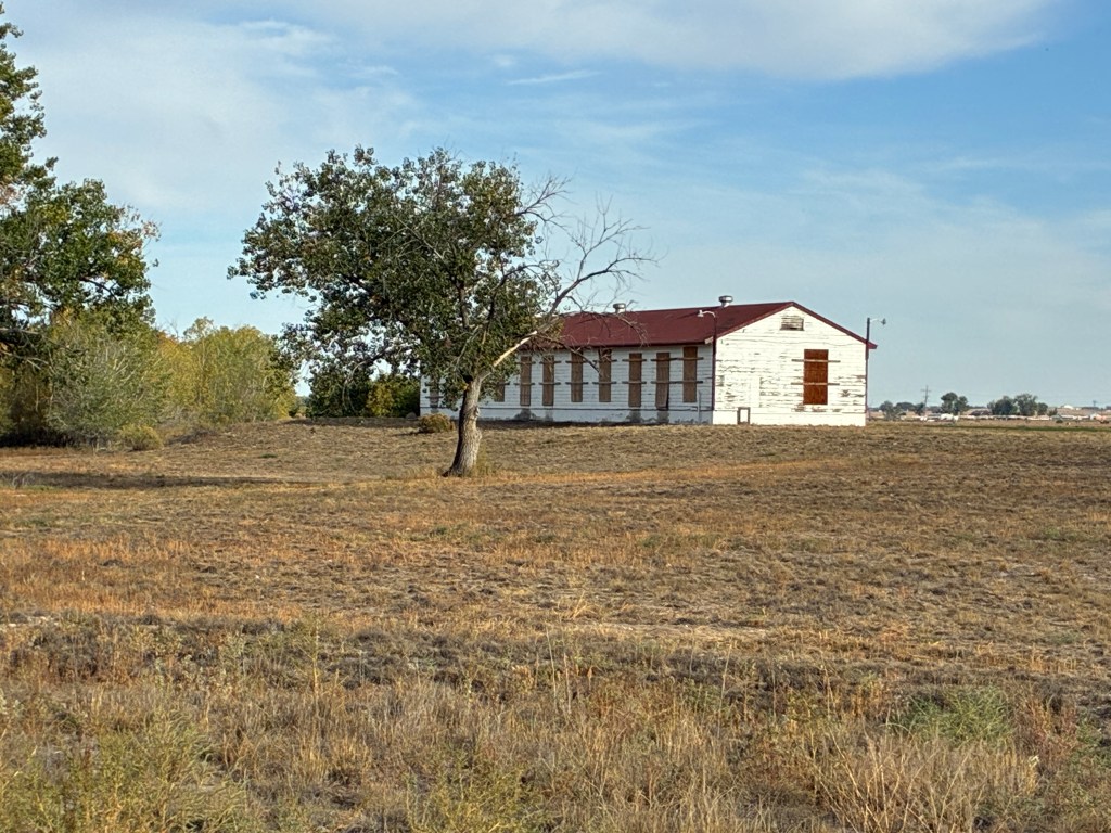 A weathered white building with a red roof is situated in an open field, surrounded by sparse trees and grass under a partly cloudy sky.