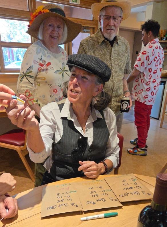 A group of people engaged in a discussion around a table with brown paper bags marked with numbers. A woman in a black hat is displaying an object, while others watch and interact in a casual setting.