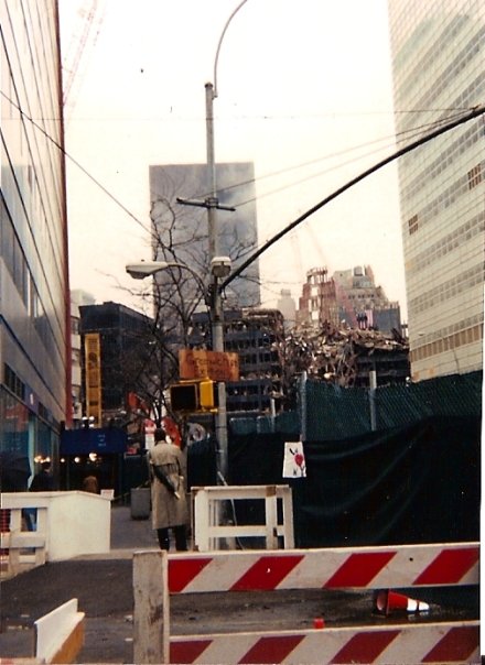 A view of a street with a construction fence and debris in the background, showing the remains of a demolished building. A person in a coat stands on the sidewalk, with other buildings and cables visible.