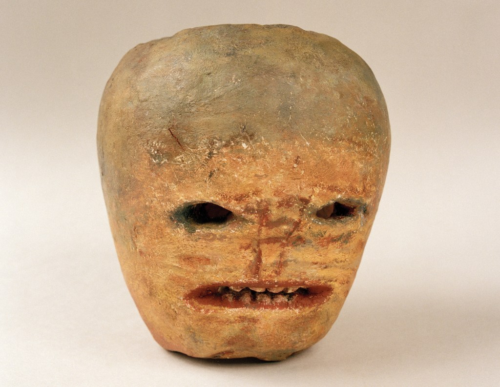 A close-up view of a sculpted head resembling a mask, with rough textures, a subtly painted face, and exaggerated facial features, including deep-set eyes and a prominent mouth.
